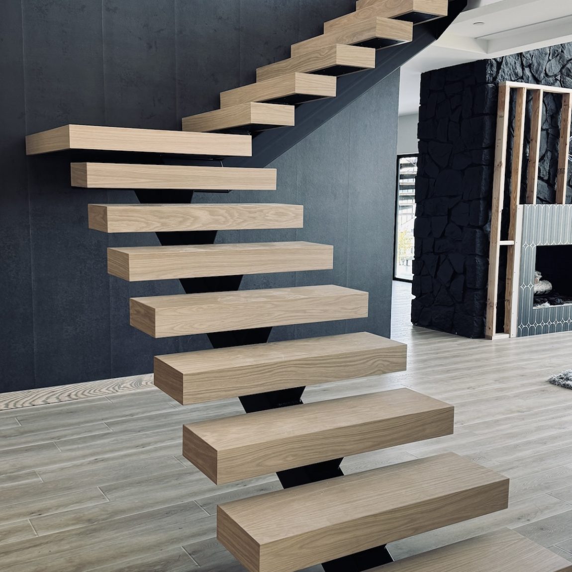 A modern cantilevered floating staircase with thick light oak wooden treads supported by hidden black metal brackets against a dark gray wall, in a contemporary open-plan interior with light wood flooring.