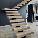 A modern cantilevered floating staircase with thick light oak wooden treads supported by hidden black metal brackets against a dark gray wall, in a contemporary open-plan interior with light wood flooring.